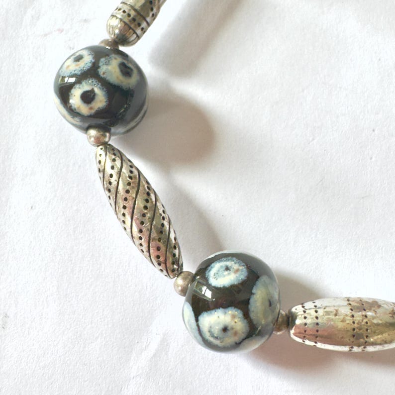 May include: Close-up of a necklace featuring large, dark beads with a floral pattern in white and light blue. The necklace also includes silver-toned, textured beads and spacers. The beads are strung together with a silver-toned chain.