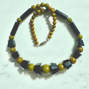 May include: A necklace featuring alternating olive green spherical beads and black geometric beads. The design includes a central large olive green sphere, flanked by black trapezoidal shapes and smaller olive green spheres, finished with a silver-toned clasp.