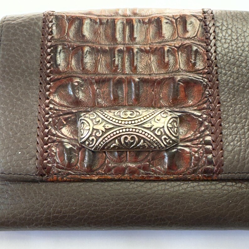 Puede incluir: Una cartera de cuero marr&oacute;n con un panel central de piel de cocodrilo sint&eacute;tica en un tono marr&oacute;n m&aacute;s oscuro. La cartera presenta un cierre plateado con un dise&ntilde;o ornamentado y detalles de cuero trenzado.
