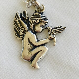 May include: Silver-toned pendant necklace featuring a detailed cherub holding a bird. The cherub is kneeling with detailed wings and a head covering. The necklace chain is delicate and silver. The pendant is approximately 1 inch in height.