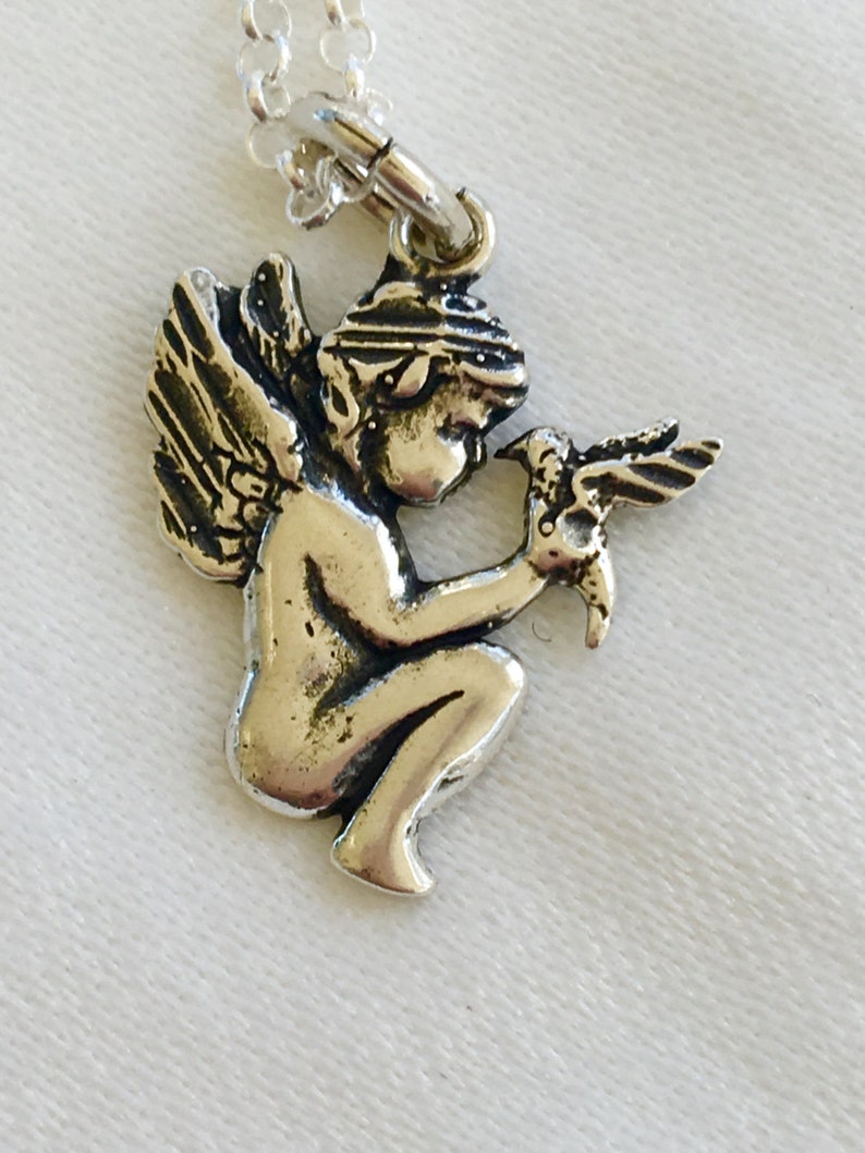 May include: Silver-toned pendant necklace featuring a detailed cherub holding a bird. The cherub is kneeling with detailed wings and a head covering. The necklace chain is delicate and silver. The pendant is approximately 1 inch in height.