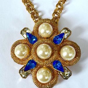 Puede incluir: Collar dorado con un colgante grande y ornamentado. El colgante presenta un grupo central de perlas de imitación, detalles azules en forma de lágrima y cristales transparentes facetados. La cadena es un diseño de eslabones grueso y texturizado.
