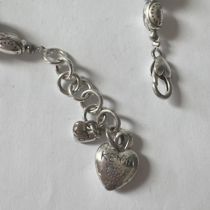 May include: Close-up of a silver-toned bracelet with a heart-shaped charm. The charm is engraved with the word "KENYA". The bracelet has a lobster clasp and a series of small, decorative beads.