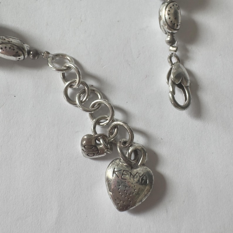 May include: Close-up of a silver-toned bracelet with a heart-shaped charm. The charm is engraved with the word "KENYA". The bracelet has a lobster clasp and a series of small, decorative beads.