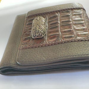 Puede incluir: Cartera plegable de cuero marr&oacute;n y beige. La parte delantera de la cartera presenta un patr&oacute;n texturizado similar al de un cocodrilo y un cierre decorativo de metal plateado. La cartera est&aacute; plegada.