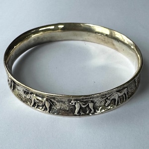 May include: A polished silver bangle bracelet featuring a textured surface with embossed animal figures including a rhinoceros, a primate, and a zebra. The interior of the band is smooth and includes the stamped text KABANA STERLING.