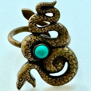 May include: A brass-toned metal ring featuring a coiled snake design with textured scales. The snake is positioned to wrap around the finger, and a small, round, bright turquoise-colored stone is set in the center of the snake's body.