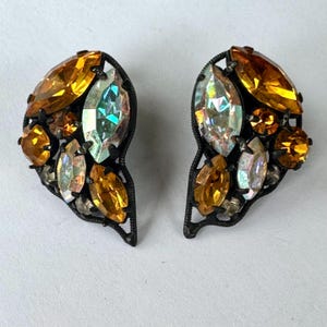 May include: A pair of vintage clip-on earrings. Each earring features a cluster of amber and iridescent crystals set in a dark metal filigree frame. The crystals are various shapes and sizes.