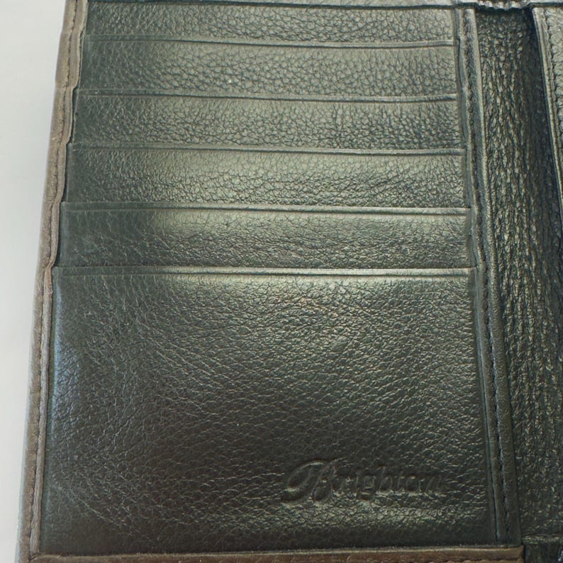 Puede incluir: Cartera de cuero negro con m&uacute;ltiples ranuras para tarjetas. La cartera tiene una superficie texturizada y un borde marr&oacute;n. El nombre de la marca "Brighton" est&aacute; grabado en la esquina inferior derecha.