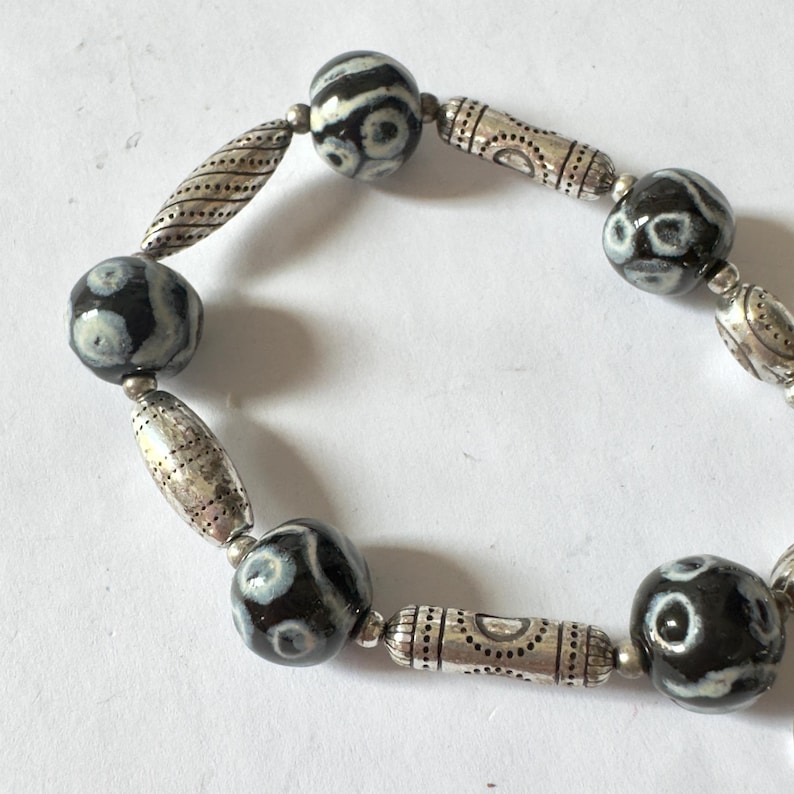 May include: A beaded bracelet featuring alternating black and white patterned beads and silver-toned metal spacers. The bracelet has a mix of round and cylindrical beads, creating a unique and stylish accessory.