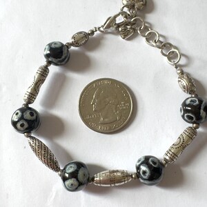 May include: A silver-toned bracelet featuring alternating black and white patterned beads and decorative silver-toned metal beads. The bracelet is shown next to a U.S. quarter for size reference. The bracelet has an adjustable chain closure.