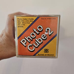 May include: A clear plastic Photo Cube 2 box with an orange and yellow label. The label reads "Photo Cube 2" in white letters and lists photo sizes. The box is held by a hand.