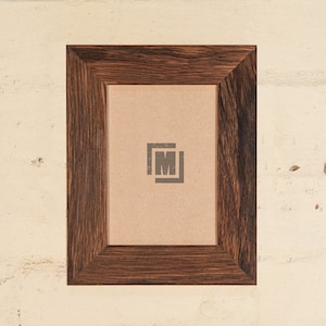 May include: A dark brown wooden picture frame with a light brown mat and a small logo in the center. The frame is approximately 8 inches by 10 inches.