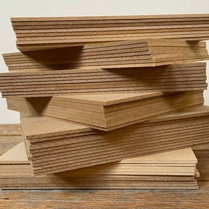 May include: A stack of unfinished, rectangular wooden boards. The boards are a light brown color and appear to be made of pressed wood or particle board. The edges of the boards show the layered construction. The boards are stacked in a slightly haphazard arrangement.