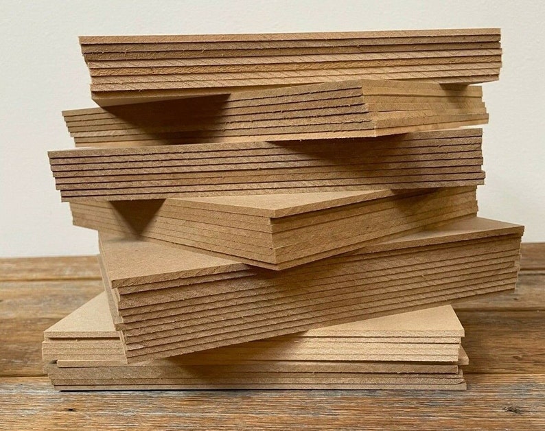 May include: A stack of unfinished, rectangular wooden boards. The boards are a light brown color and appear to be made of pressed wood or particle board. The edges of the boards show the layered construction. The boards are stacked in a slightly haphazard arrangement.