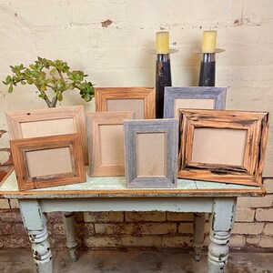 May include: Six rustic wooden picture frames of various sizes and colours, including brown, grey, and natural wood, arranged on a light green table with a brick wall in the background.
