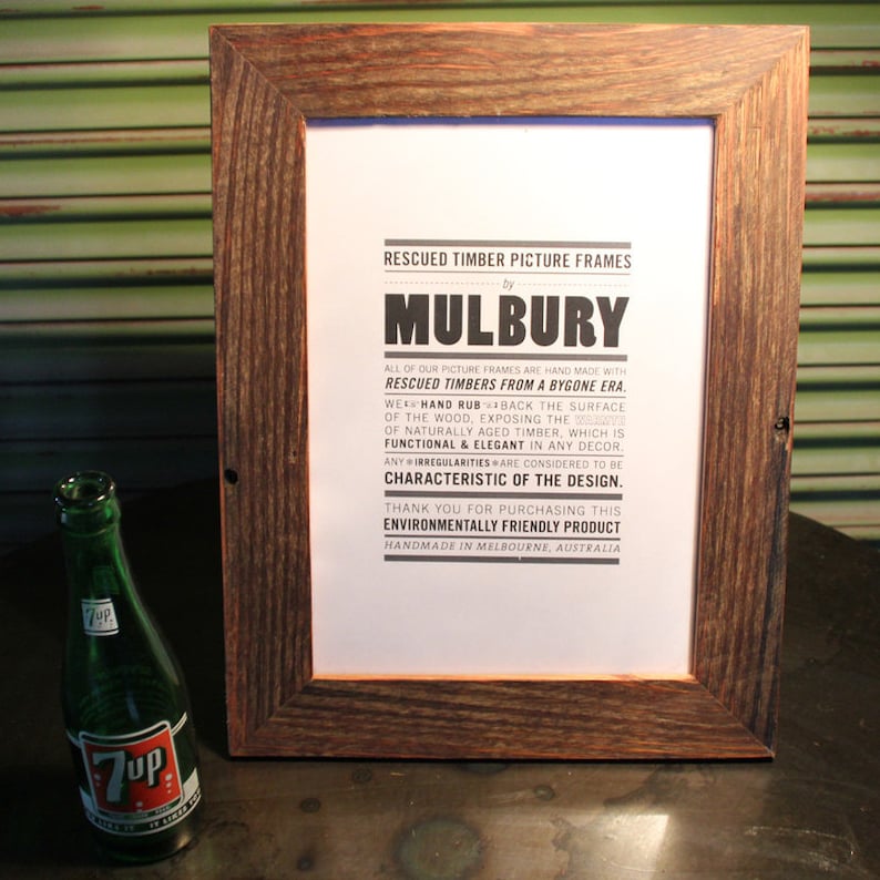May include: A brown wooden picture frame with a white paper insert. The paper has black text that reads "Rescued Timber Picture Frames by Mulbury. All of our picture frames are hand made with rescued timbers from a bygone era. We hand rub back the surface of the wood, exposing the ... characteristic of the design. Thank you for purchasing this environmentally friendly product. Handmade in Melbourne, Australia."
