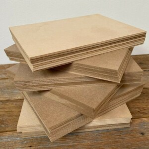 May include: A stack of light brown rectangular wooden boards. The boards are arranged in a staggered pile, showing their clean edges and uniform size. The wood has a natural, unfinished appearance.