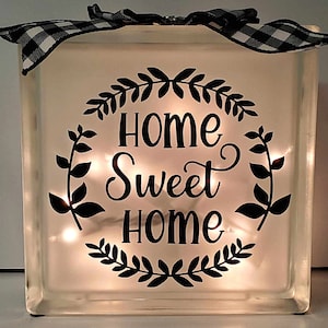 May include: A white glass block with a black and white checkered ribbon on top. The block has a black vinyl decal with a wreath of leaves and the text "Home Sweet Home". The block is illuminated with white LED lights.