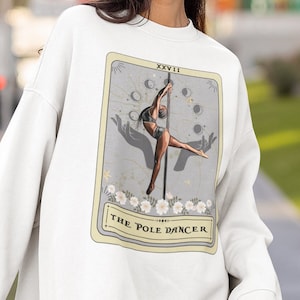 Könnte beinhalten: Weißer Pullover mit einem Grafikdruck einer Tarotkarte, auf der eine Frau am Poledance zu sehen ist. Die Karte ist mit "The Pole Dancer" und "XXVIII" beschriftet.