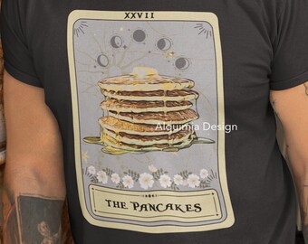May include: A black t-shirt with a graphic of a tarot card featuring a stack of pancakes with a yellow and brown color scheme. The card is labeled "The Pancakes" and "XXVII" in a vintage style font.