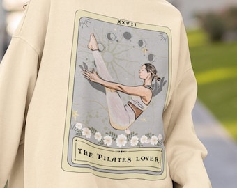 May include: A cream-colored sweatshirt with a graphic of a tarot card featuring a woman doing a pilates pose. The card is labeled "XXVIII The Pilates Lover".