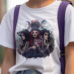 Puede incluir: Camiseta blanca con un estampado gr&aacute;fico de tres brujas con una luna creciente y estrellas. Las brujas llevan sombreros y t&uacute;nicas negras y sostienen varios objetos, entre ellos una caldera, una varita y un libro.