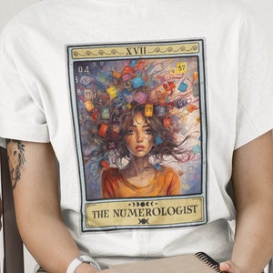 May include: White t-shirt with a colorful illustration of a woman with numbers in her hair. The illustration is framed with a border that says "XVII THE NUMEROLOGIST".