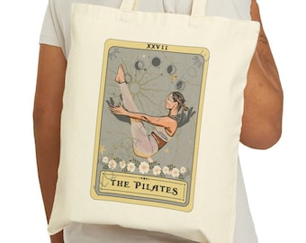 Puede incluir: Una bolsa de tela beige con un diseño de carta de tarot amarilla y marrón. La carta presenta a una mujer haciendo un ejercicio de pilates con el texto "The Pilates" y "XXXVII" en la carta.