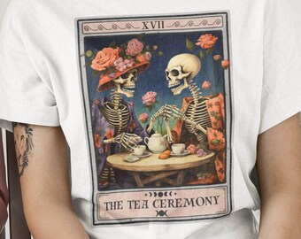 Puede incluir: Camiseta blanca con un gráfico de dos esqueletos tomando el té. El gráfico es de estilo vintage con un fondo azul y detalles florales. El texto "The Tea Ceremony" está debajo del gráfico.