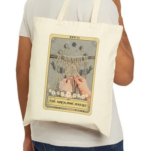 May include: A beige tote bag with a cream canvas design featuring a tarot card illustration. The card is labeled "XXVII" and "THE MACRAME ARTIST".