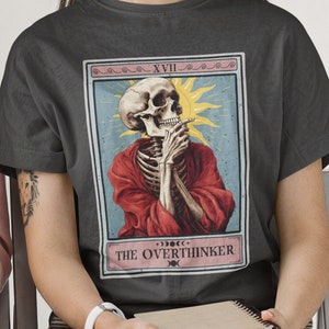 May include: A dark gray t-shirt with a tarot card design featuring a skeleton in a red robe, a sun, and the text "XVII THE OVERTHINKER".