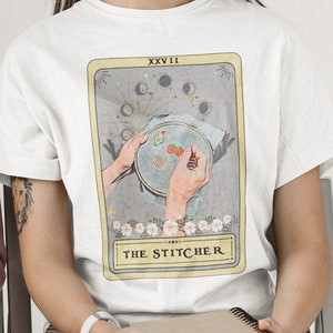 May include: White t-shirt featuring a graphic of a tarot card. The card is titled "The Stitcher" and depicts a hand holding a needle and thread. The card is framed in gold and has a moon and star design.