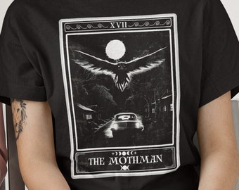 May include: Black t-shirt with a white graphic of a tarot card depicting a creature with large wings flying over a car on a dark road. The card is labeled "XVII" and "THE MOTHMAN".