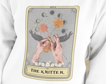 May include: White sweatshirt with a graphic of a tarot card featuring a pair of hands knitting with yarn and the text "THE KNITTER" on a yellow background.