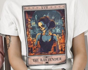 May include: White t-shirt with a tarot card design featuring a woman with a skull face behind a bar with bottles. The card is labeled "XVII THE BARTENDER".