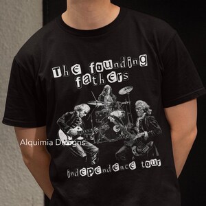 Może przedstawiać: Czarny T-shirt z grafiką przedstawiającą czterech mężczyzn grających na instrumentach. Na koszulce nadrukowany jest tekst "The founding fathers independence tour". Na koszulce nadrukowany jest również tekst "Alquimia Designs".
