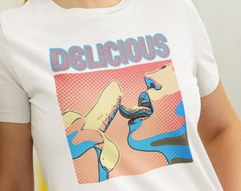 Peut inclure: T-shirt blanc avec un graphisme de style pop art. Le graphisme représente une banane en train d'être mangée, avec le mot "DELICIOUS" au-dessus. Le design utilise des couleurs rouge, bleu et jaune.