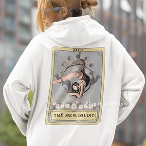 May include: White hoodie featuring a tarot card design depicting a woman performing aerial acrobatics in a hoop. The card is labelled "The Aerialist" and bears the number XXVIII. The design is in a vintage style with a grey background and floral details.