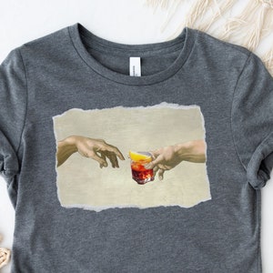 May include: A dark gray t-shirt with a print of two hands reaching towards each other, holding a glass of red liquid with a lemon wedge. The print is inspired by the famous Renaissance painting "The Creation of Adam".