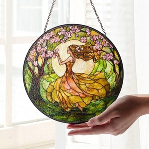 Op de afbeelding: Een gebrandschilderd raam dat aan een ketting hangt. Het raam toont een vrouw in een vloeiende gele jurk die in een bos staat met een boom en roze bloemen.