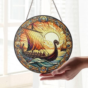 Puede incluir: Una ventana de vitral colgada de una cadena. La ventana representa un barco vikingo navegando en un mar al atardecer con un sol dorado y olas azules. El barco tiene una vela marrón y un diseño rojo y dorado.