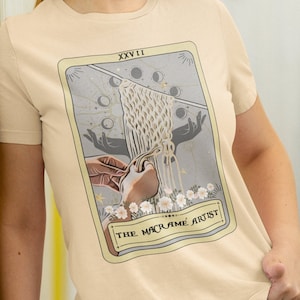 Op de afbeelding: Een beige t-shirt met een afbeelding van een tarotkaart. De kaart heeft de titel "De macramé-kunstenaar" en toont een hand die een macraméknoop vasthoudt tegen een achtergrond van een halve maan en sterren.