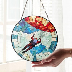 Könnte beinhalten: Ein Buntglasfenster, das an einer Kette hängt. Das Fenster ist rund und zeigt einen Fallschirmspringer mit einem rot-weiß-blauen Fallschirm. Der Fallschirmspringer fällt durch einen blauen Himmel mit weißen Wolken.