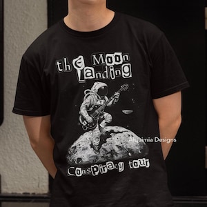 May include: Black t-shirt with a graphic of an astronaut playing a guitar on the moon. The text "the Moon Landing Conspiracy tour" is printed on the shirt.
