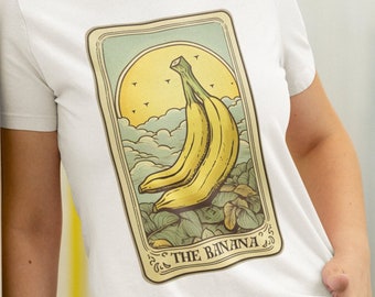 Peut inclure: T-shirt blanc avec un motif de carte de tarot représentant une banane jaune. La carte a une bordure vert clair et les mots "THE BANANA". Le fond comprend un soleil jaune, des nuages et des oiseaux.