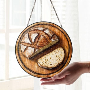 Puede incluir: Un letrero redondo de madera con una cadena de metal para colgar. El letrero presenta una imagen pintada de una hogaza de pan y una rebanada de pan.