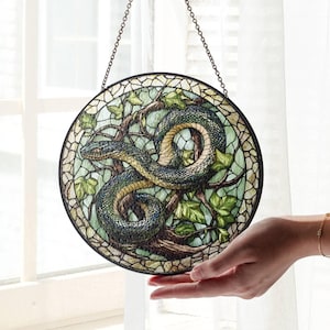 Op de afbeelding: Een gebrandschilderd raam dat aan een ketting hangt. Het raam is rond en toont een groene slang die om een ​​boomtak met groene bladeren is gewikkeld. De achtergrond is lichtblauw.
