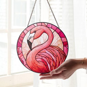 Puede incluir: Una ventana de vidrio coloreado que cuelga con un diseño de flamenco rosa. El flamenco está de perfil con la cabeza girada hacia la izquierda. El fondo es una mezcla de rosa, naranja y amarillo.