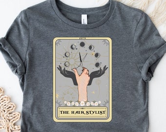 May include: A grey t-shirt with a graphic of a tarot card design featuring a hand holding scissors and the text "THE HAIR STYLIST".